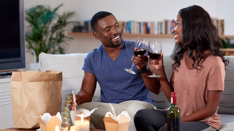 A couple sitting on the couch enjoying a glass of wine and takeout food.