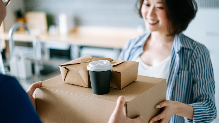 Woman receiving her delivery takeout order (two brown boxes and a drink in a to-go cup).