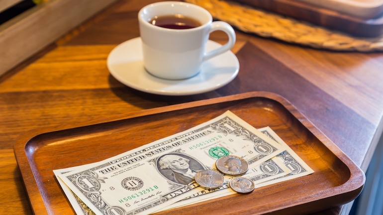 Dollar bills and coins on a wooden tray next to a white cup of tea or coffee on a coffee table.